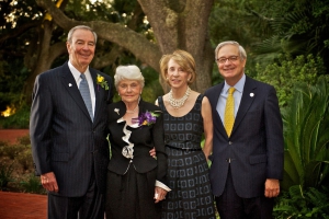From left to right, a man wearing a black suit and blue tie smiling, a woman with short white hire wearing a black dress smiling, a woman with short brown hair wearing a black dress and pearls smiling, and a man wearing a blue suit with a yellow tie and glasses smiling.
