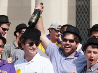 A male student wearing a black hat smiles and holds up a bottle of champagne