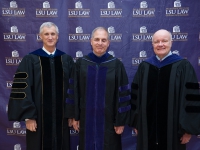 A group of people wearing graduation robes and caps smile with the LSU Law logo in the background