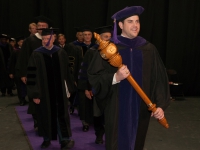 A male student wearing graduation attire carries a wooden mace