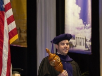 A male student wearing graduation attire carries a wooden mace