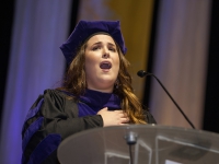 A female student wearing graduation attire places her hand over her heart and sings
