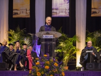 A man wearing graduation attire speaks at a podium