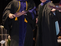 A male student wearing graduation attire and holding a diploma walks off the stage