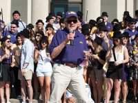A man  wearing an LSU hat holds a microphone and talks while a group of students is in the background