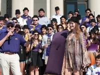 A man  wearing an LSU hat holds a microphone and talks while a group of students is in the background
