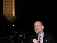 A man talks in a microphone with the Louisiana state capitol in the background.
