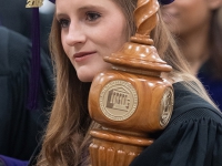 A female student wearing graduation attire carries a wooden mace