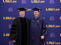 Two men wearing graduation robes and caps smile with the LSU Law logo in the background