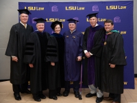 A group of people wearing graduation robes and caps smile with the LSU Law logo in the background
