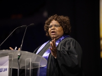 A woman wearing graduation attire speaks at a podium