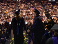 A female student wearing graduation attire walks across the stage