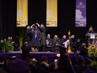 A female student smiles as two men put a graduation robe over her