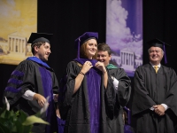 A female student smiles as two men put a graduation robe over her