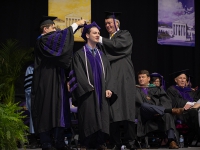 A male student smiles as two men put a graduation robe over her