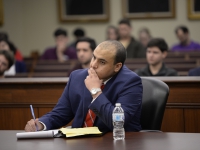 A male student wearing a suit and tie sits at a table and takes notes