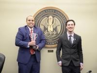 A male student wearing a suit and tie holds a crystal trophy and smiles next to the LSU Law Center seal and another male student wearing a suit and tie