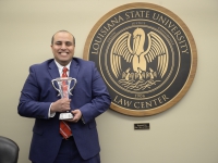 A male student wearing a suit and tie holds a crystal trophy and smiles next to the LSU Law Center seal