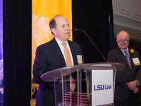 A man wearing a suit and tie talks at a podium with purple and gold banners in the background