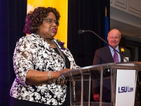 A woman talks at a podium with purple and gold banners in the background