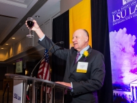 A man wearing a suit and tie holds up a glass with purple and gold banners in the background