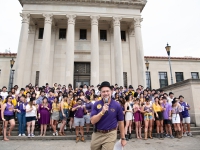A man talks on a microphone with a group of people behind him on the steps of the LSU Law Center