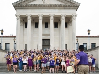 Students  wearing derby hats lift up a champagne glass and smile with the LSU Law Center in the background