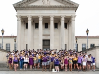 A group of students  wearing derby hats lift up champagne glasses and smile with the LSU Law Center in the background