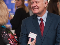 A man wearing a suit and tie smiles as he talks to a woman