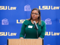 A female student wearing a green dress speaks at a podium with the LSU Law logo in the background