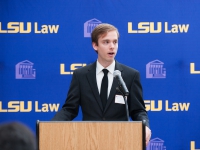 A male student wearing a suit and tie speaks at a podium with the LSU Law logo in the background