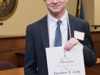 A male student wearing business attire and white cords smiles and holds a certificate