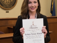 A female student wearing business attire and white cords smiles and holds a certificate