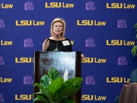 A woman in a black dress talks at a podium with the LSU Law logo in the background