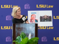 A woman in a black dress holds up a sign with the LSU Law logo in the background