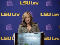 A woman wearing a brown jacket talks at a podium with the LSU Law logo in the background