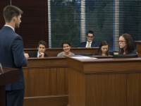 A male student wearing a suit and tie stands and talks to a seated student with a group of students in the background