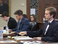 Three male students wearing suits and ties sit at a table and take notes