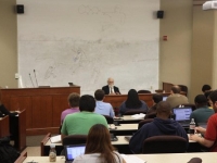 A male professor sits at the front of a classroom while students listen