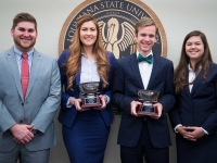 A male and female student hold silver bowl trophies and smile for a photo with another male and female student next to the LSU Law Center seal