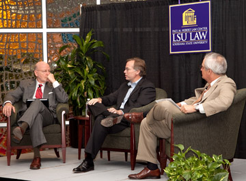 Three men in suits sit in chairs and talk with plants and stained glass in the background