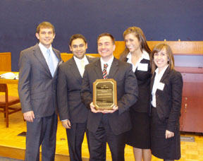 Three men and two women in suits pose for a photo with the male in the center holding a plaque