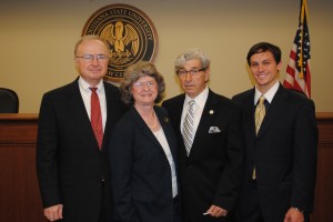 Three men and a woman pose for a photo in the LSU Law Courtroom with the state seal of Louisiana in the background