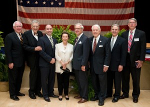 Seven men and one woman wearing suits pose for a photo with an American flag in the background