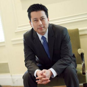 A man wearing a suit and tie sits in a chair and poses for a photo