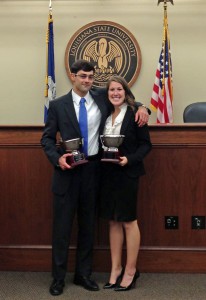 A male and female student wearing suits and holding trophies smile for a photo with the LSU Law courtroom in the background