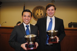 Two male students wearing suits hold trophies and smile for a photo with the LSU Law courtroom in the background