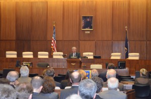 A man speaks at a podium with a wooden wall in the background