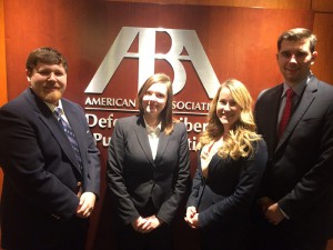 Two men and two women wearing suits pose for a photo with the American Bar Associaion logo on a wooden wall in the background