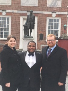 Two men and a woman wearing suits pose for a photo with a statue in the background